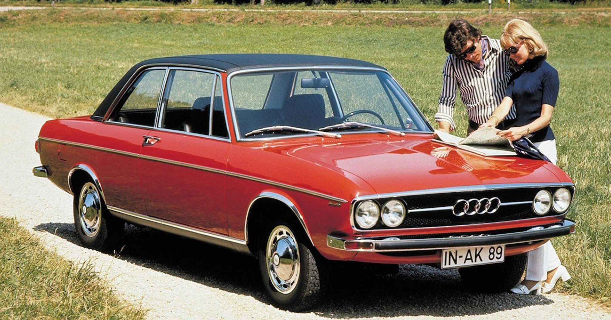 A side view of a red 1968 Audi 100 Coupe with two models consulting an old map on the hood.