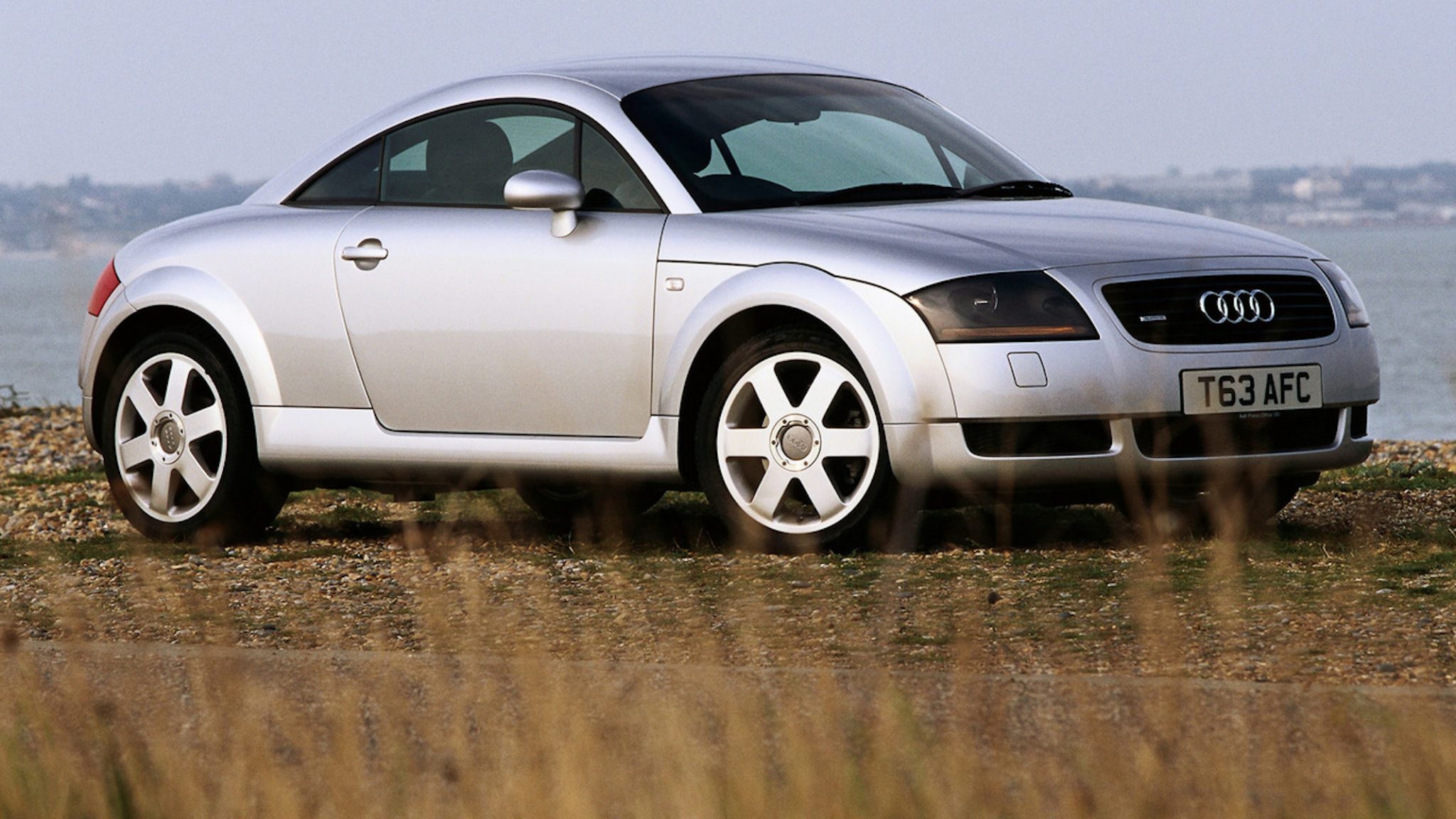 A side view of a silver First Gen Audi TT Coupe in front of a large body of water.