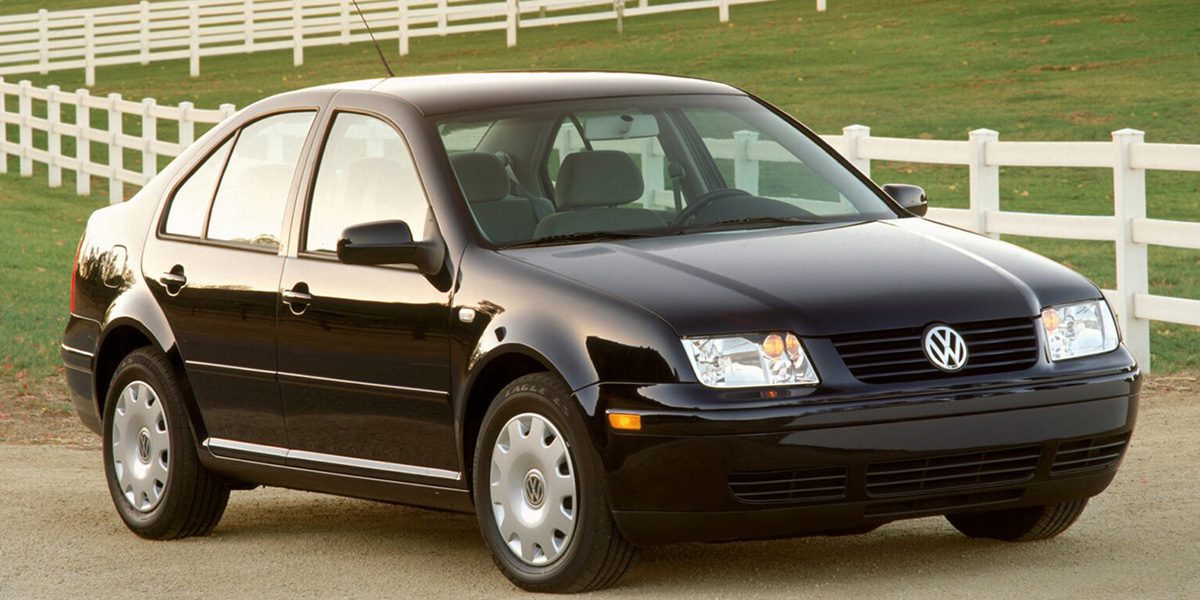 A side view of a Black four-door MK4 VW Jetta on a dirt road in front of a white picket fence