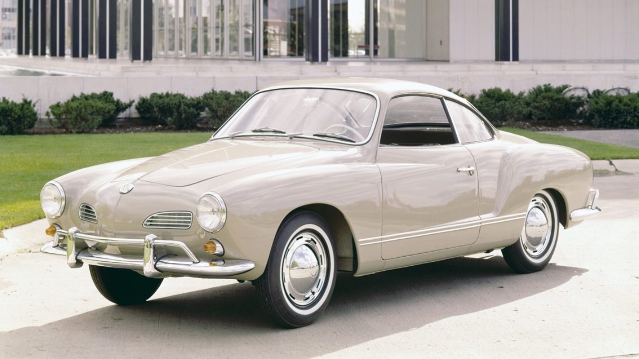 A side view of a 1950s beige VW Karmann-Ghia on white pavement in front of a house