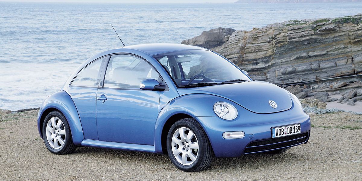 A side view of a 1990s blue VW new Beetle parked on the beach next to a body of water